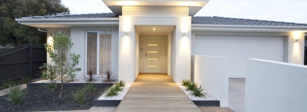 Bílý moderní rodinný dům s dřevěným chodníkem a rozsvíceným nástěnným venkovním osvětlením